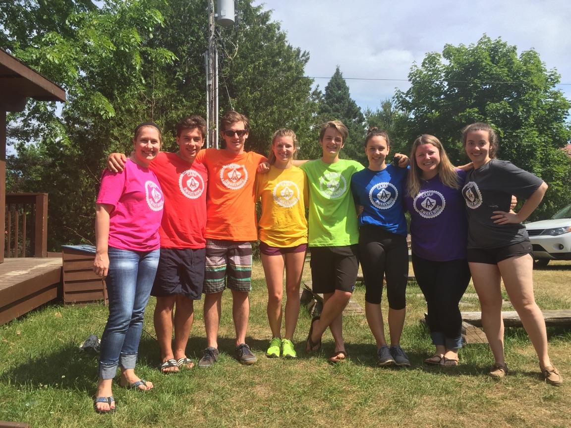 Group photo of Camp Awakening staff members wearing a rainbow of coloured t-shirts with the camp logo