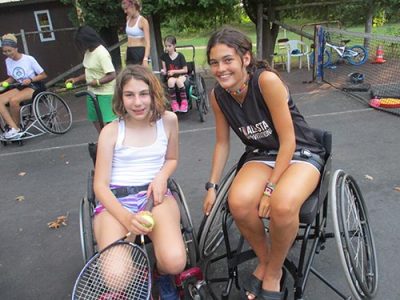 Smiling camper and staff member posing in sport wheelchairs during a game of tennis